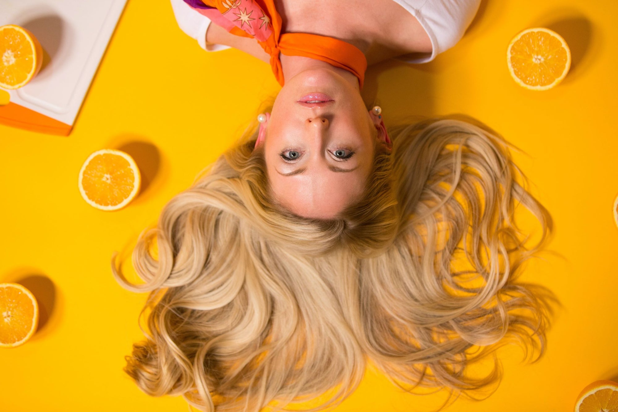 Mulher deitada com cabelos loiros espalhados em fundo amarelo com pedaços de laranja ao redor.